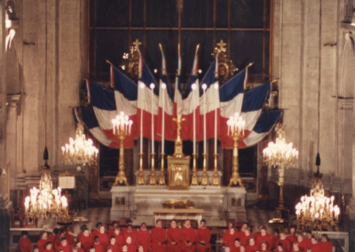 A choir dressed in red robes stands in a semicircle in a grand church with chandeliers, a gold altar, and large French flags displayed behind them. A conductor leads the choir on a checkered marble floor.