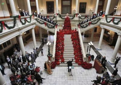 A large choir dressed in red sings on a grand staircase in a decorated hall, surrounded by an audience and festive garlands, likely during a holiday event or ceremony.