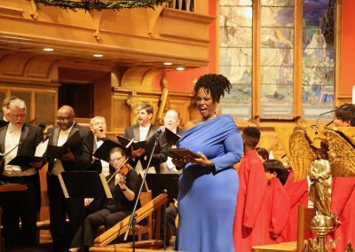 A woman in a bright blue dress sings in front of a choir and musicians inside a church with stained glass windows and wooden interior. Several choir members wear black and some are dressed in red robes.