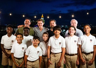 A group of boys in white polos and khaki shorts stand in front of four adults at night, smiling outdoors with city lights and string lights in the background.