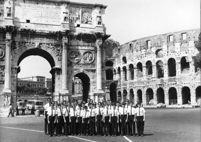 A group of people in uniform pose in front of the Arch of Constantine and the Colosseum in Rome on a sunny day, with vintage cars and buildings visible in the background.