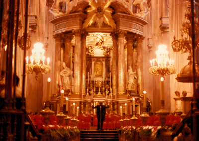 A conductor stands on stage inside an ornate, golden-lit church or cathedral with grand columns, chandeliers, and red seats, facing an altar adorned with statues and religious decorations.