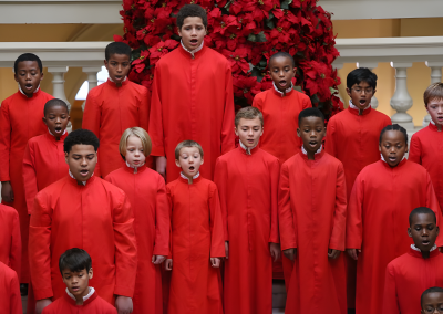 A choir of boys wearing matching red robes sings in front of a large display of red poinsettias, standing in rows on indoor steps with white railings.