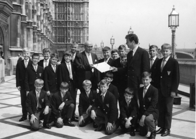 A group of boys in blazers pose outside a historic building with two men in suits, one holding a document. The setting features ornate architecture and street lamps, likely near the Houses of Parliament in London.