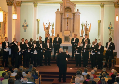 A choir of men dressed in black tuxedos sings in front of an altar inside a church, directed by a conductor. An audience is seated, watching the performance. The church features ornate decorations and religious statues.