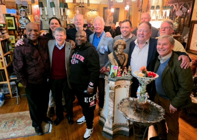 A group of eleven smiling men pose together indoors in a warmly decorated room with wooden floors, ornate furniture, and a bust sculpture surrounded by flowers in the center.