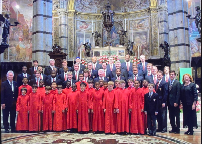 A large choir group, including boys in matching red robes and adults in suits, poses for a photo inside an ornate church with marble columns and detailed religious artwork.