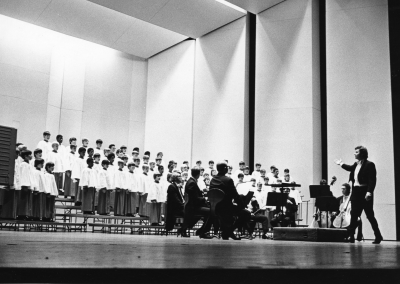 A black and white photo of a choir of children in robes performing on stage with an orchestra. A conductor stands in front, directing both the choir and musicians. The background features tall, geometric acoustic panels.