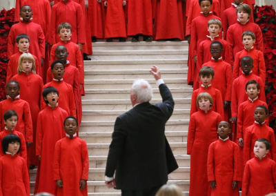 A choir of children in matching red robes sings on a grand staircase decorated with red wreaths, led by a conductor with white hair, while an audience watches from below.