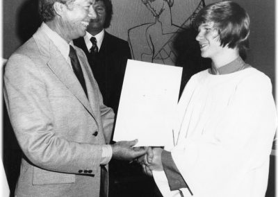 A man in a suit smiles and shakes hands with a young person in a choir robe, as they present a document. Two other people stand in the background in front of abstract art. The photo is in black and white.