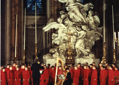 A choir dressed in red robes performs in a grand church with stained glass windows and a large white religious statue behind them. A harpist is positioned in front, and the conductor stands facing the choir.
