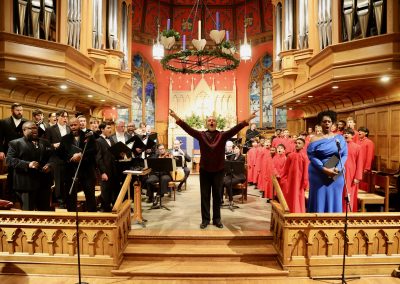 A joyous choir and orchestra perform in a grand church with stained glass windows. The conductor stands at the center with raised arms, while singers in black and red robes and a soloist in blue stand around him.