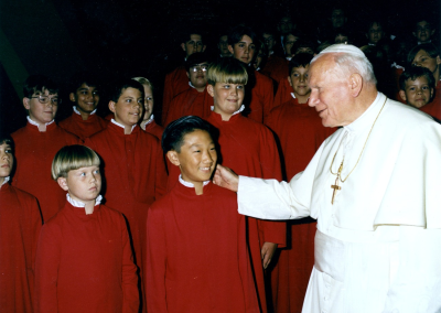 A man in white religious robes and a cross warmly greets smiling children in matching red choir gowns, standing together in several rows.