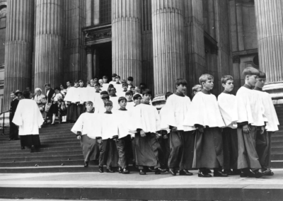 A group of choir boys in white robes walk down the steps of a large building with tall columns, followed by adults and more children.